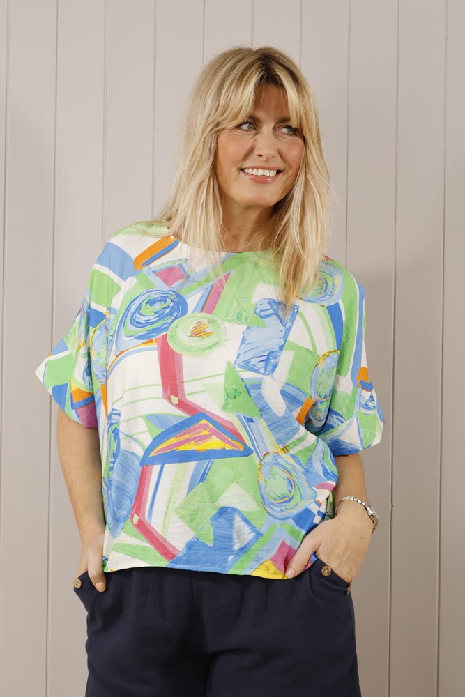 A woman wearing a multicolored abstract print scoop neck top with batwing sleeves, paired with white trousers.