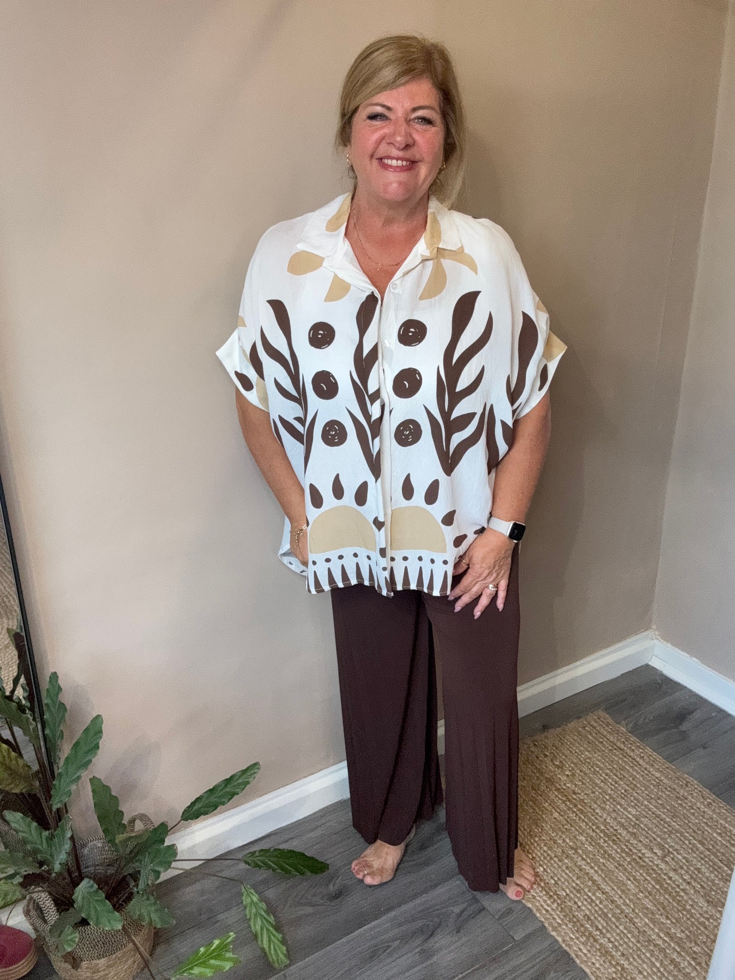 Woman standing indoors wearing a patterned top and brown pants.