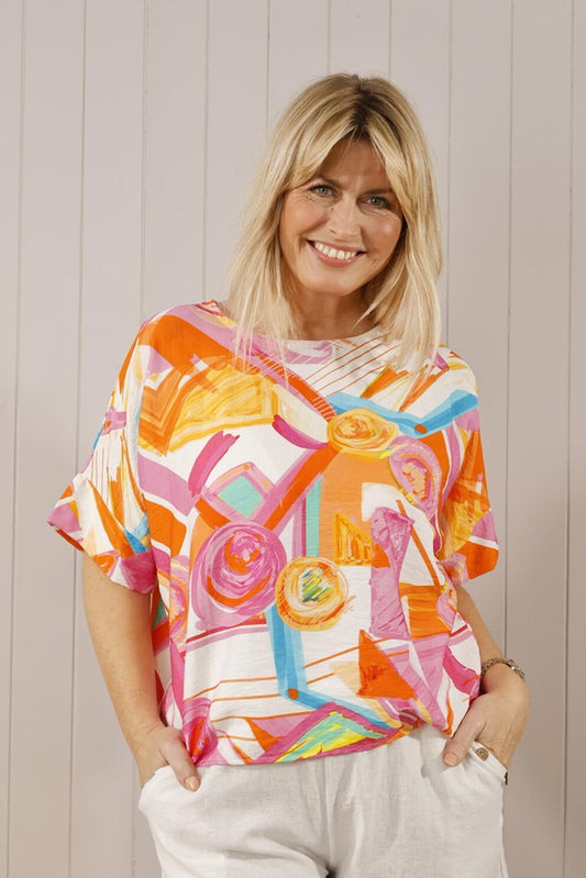 A woman wearing a multicolored abstract print scoop neck top with batwing sleeves, paired with white trousers.