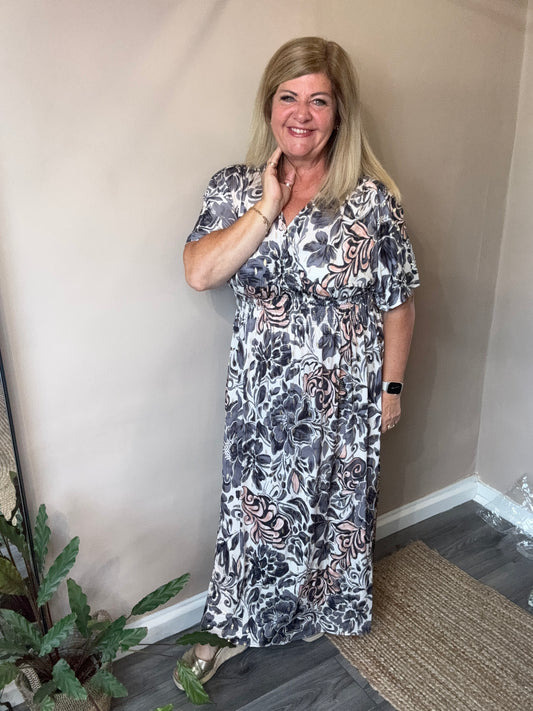 Woman wearing a floral dress standing indoors with a plant in the foreground.